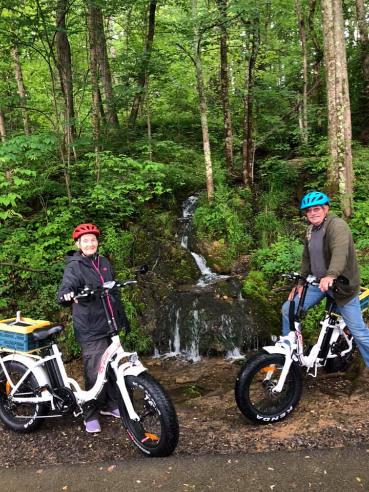 Two smiling customers posing with two DJ Folding Bike Step Thru step-through fat tire e-bikes on a dirt path in a lush green forest