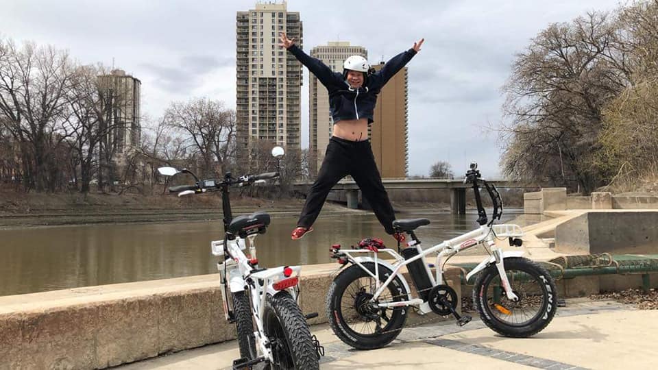A happy customer jumping for joy with two DJ Folding Bike fat tire e-bikes on a paved path in front of a city skyline
