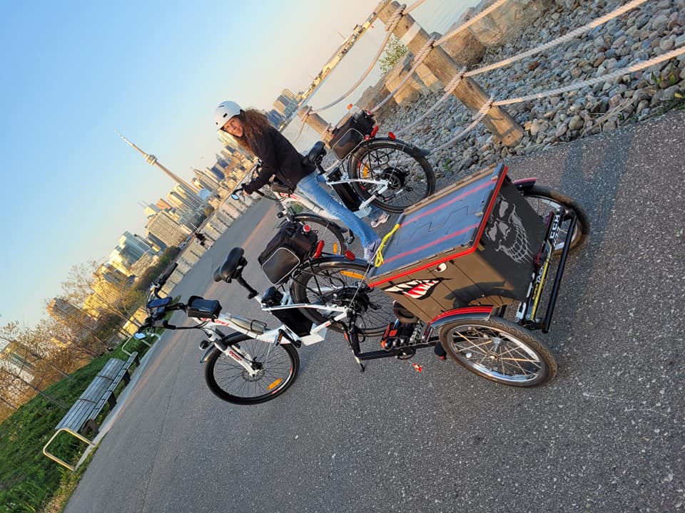 A smiling rider poses on a paved waterfront path with two DJ City Bike e-bikes equipped with trunk bags, cell phone holders, and rear trailer