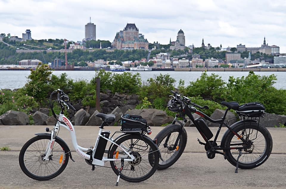 A DJ City Bike e-bike and a DJ Fat Bike Mid Drive fat tire e-bike on a paved road in front of the St. Lawrence River and Québec City