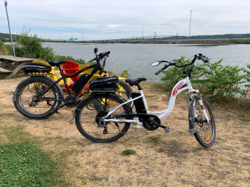 A DJ Fat Bike electric bike equipped with trunk bag and front camera, and a DJ City Bike electric bike equipped with trunk bag and rear view mirror on a grassy beach in front of a body of water