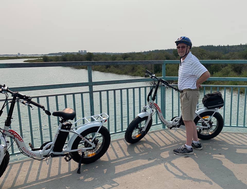 A smiling customer posing with two DJ Folding Step Thru fat tire e-bikes in front of the Glenmore Reservoir in Calgary, Alberta