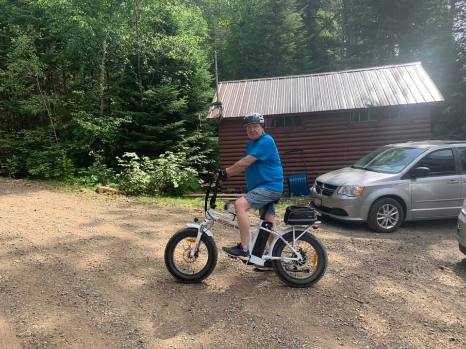 A smiling customer riding a DJ Folding Bike fat tire e-bike equipped with pannier bag on a dirt road in front of a cabin in a forested area